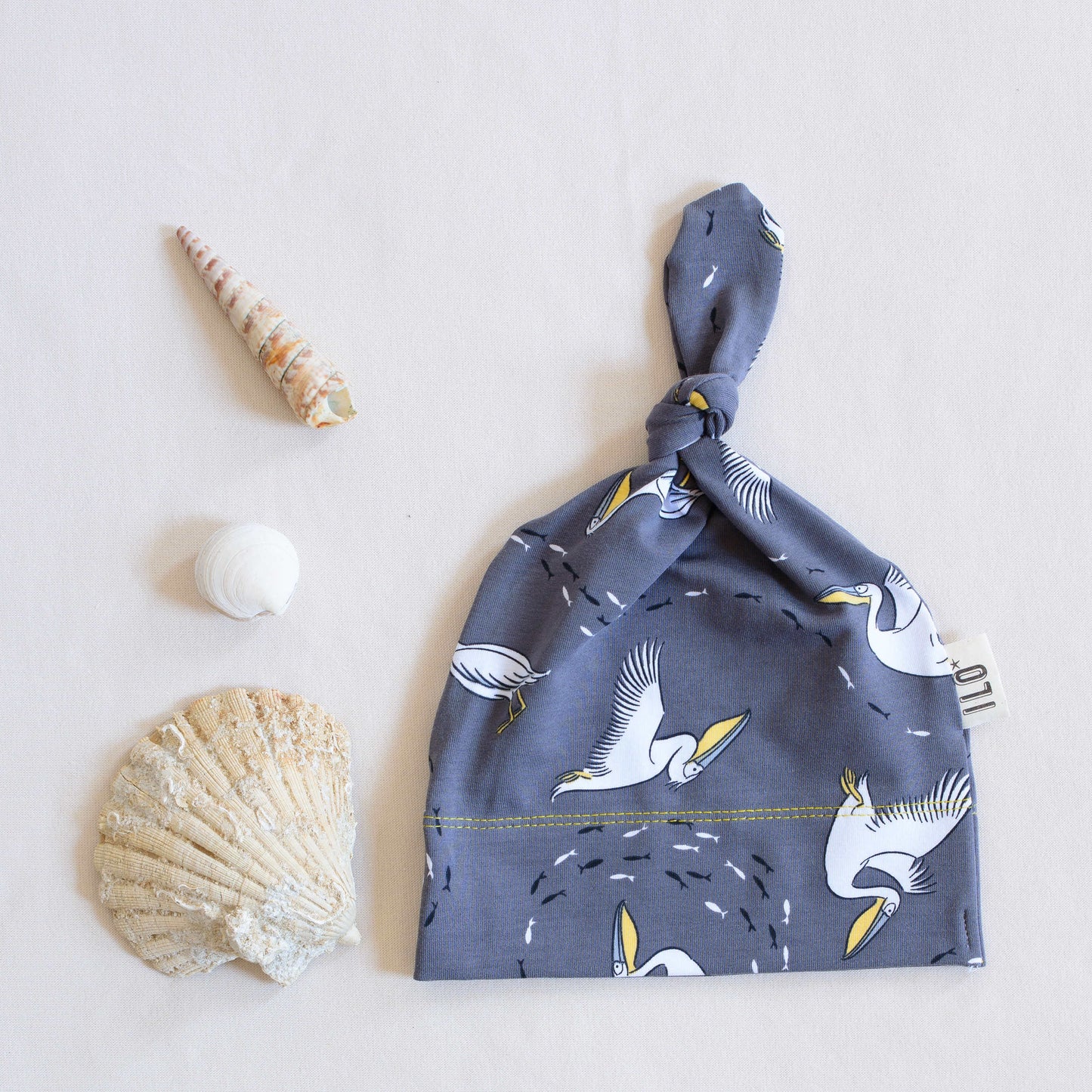 Pelicans organic knot hat with sea shells by it on a soft cream background