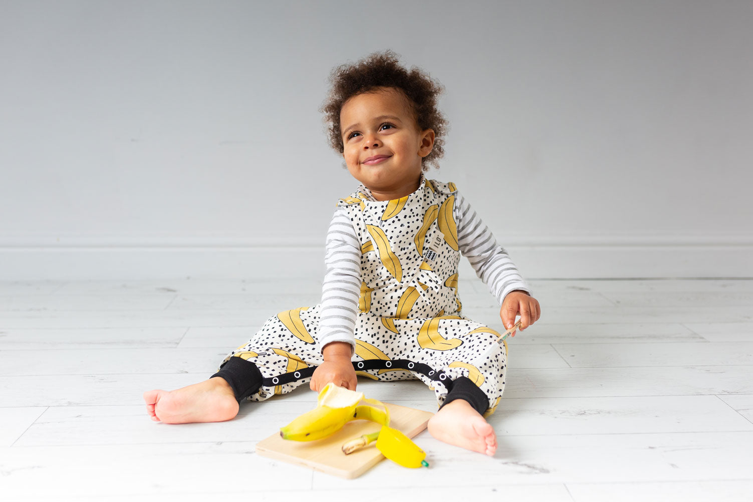 young child wearing banana print romper playing with bananas in a white room ILO Clothing
