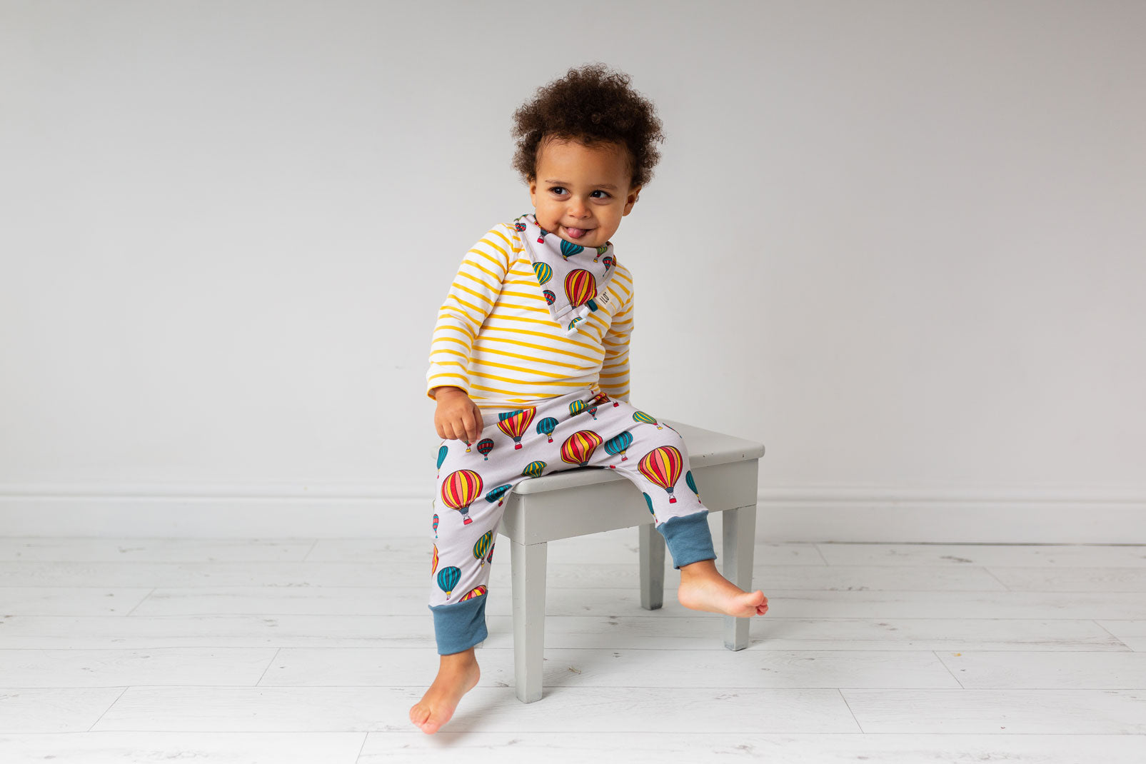 young child wearing balloons print stone colour trousers and bib sat on a chair in a white room ILO Clothing